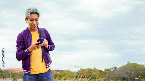 hombre joven moreno sonriente con celular en mano y audífonos ropa morada y amarilla con pelo pintado feliz 