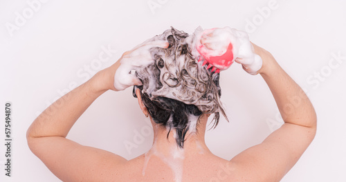 Lady with foam in her hair takes care of her scalp with pink massager against white background.
