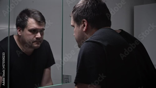 Upset lonely man suffering from depression looking in reflection at bathroom mirror. Concept of depression, suicide, stress, mental illness, loneliness and frustration
