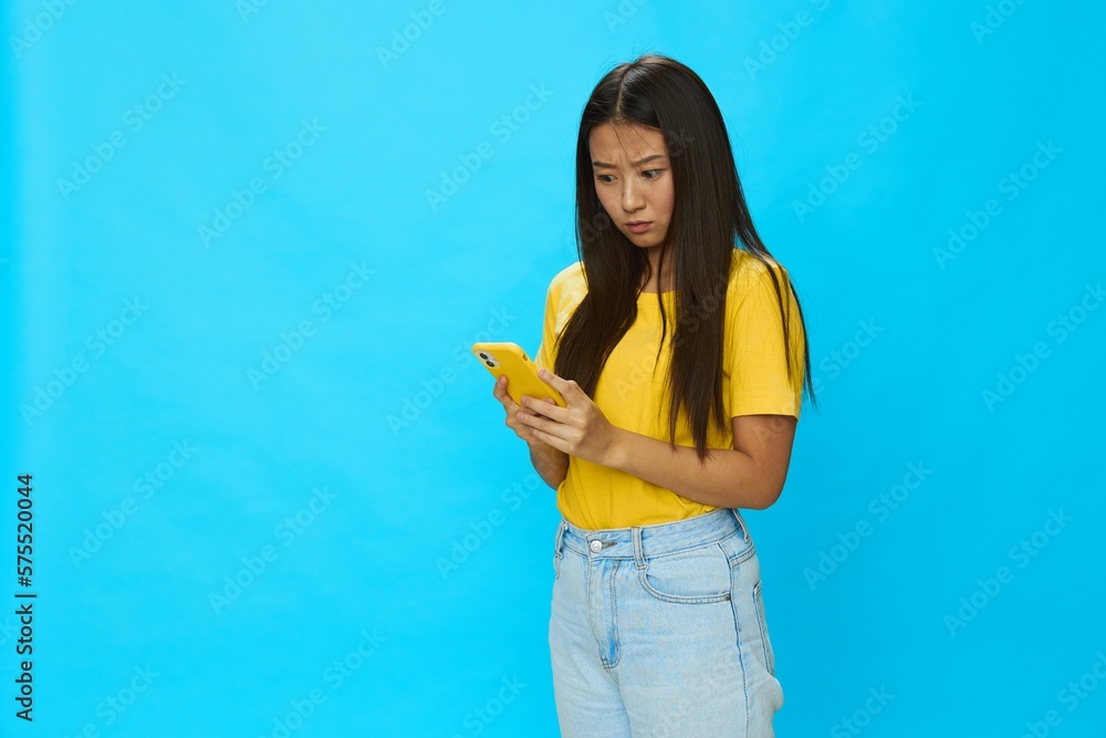 Happy Asian woman holding a phone and looking at the screen with a yellow case on a blue background in a yellow T-shirt smiling with teeth Happy Asian woman holding a phone and looking at the screen with a yellow case on a blue background in a yellow T-shirt smiling with teeth