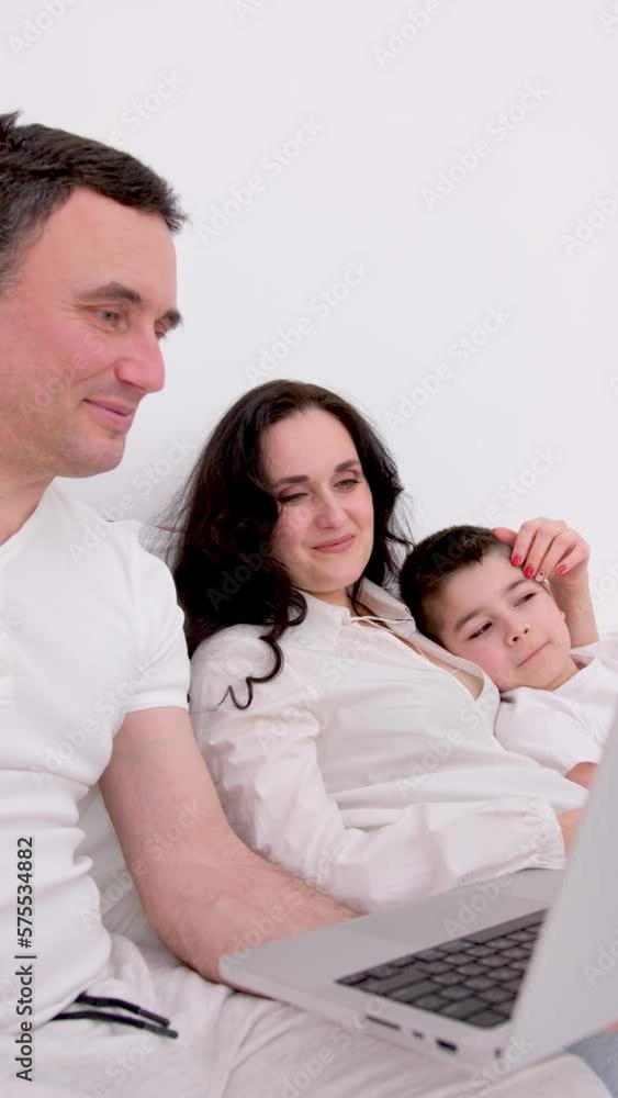 family in bed playing on tablet sincerely laughing having fun mom father and son dark hair white big duvet family is watching movies with the son on laptop good way to spend time on quarantine. family in bed playing on tablet sincerely laughing having fun mom father and son dark hair white big duvet family is watching movies with the son on laptop good way to spend time on quarantine.