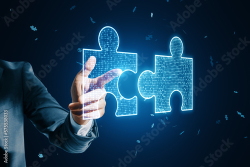 Close up of male hand pointing at glowing digital blue jigsaw puzzle hologram on dark blurry background. Digital solution, collaboration, partners cooperate, implement merge, matching concept.