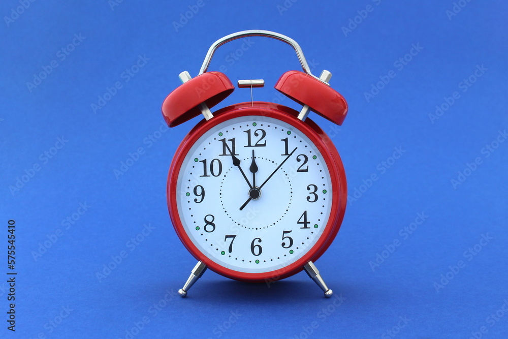 A red alarm clock stands on a blue background. The time shows five minutes to twelve. A red alarm clock stands on a blue background. The time shows five minutes to twelve.