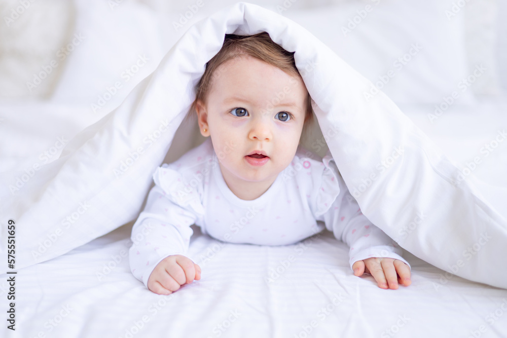 funny happy baby girl looks out from under the blanket on the bed in white clothes and smiles, a small child on a cotton bed at home woke up in the morning after sleeping funny happy baby girl looks out from under the blanket on the bed in white clothes and smiles, a small child on a cotton bed at home woke up in the morning after sleeping