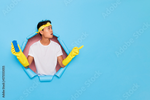 Horizontal shot of serious ethnic young man in protective yellow gloves and headband keeps cleaning brush points index finger right to copy space shows some promotion has surprised expression isolated photo