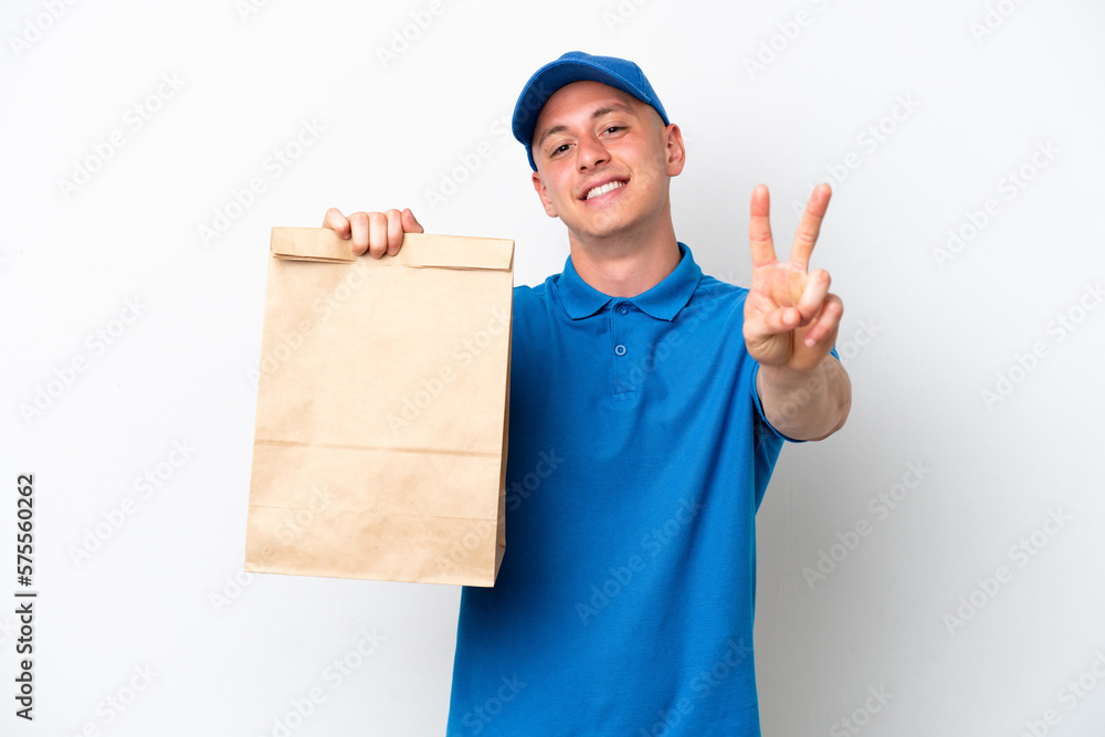 Young Brazilian man taking a bag of takeaway food isolated on white background smiling and showing victory sign Young Brazilian man taking a bag of takeaway food isolated on white background smiling and showing victory sign