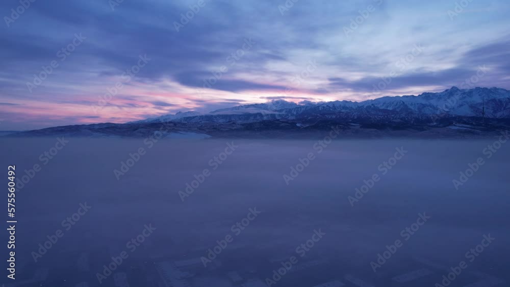 Dawn over the city in fog and smog. Mountain view. A light haze hangs over the city, cars drive, signs glow. Clouds, sun rays and snowy mountains are visible in the distance. Almaty, Kazakhstan Dawn over the city in fog and smog. Mountain view. A light haze hangs over the city, cars drive, signs glow. Clouds, sun rays and snowy mountains are visible in the distance. Almaty, Kazakhstan