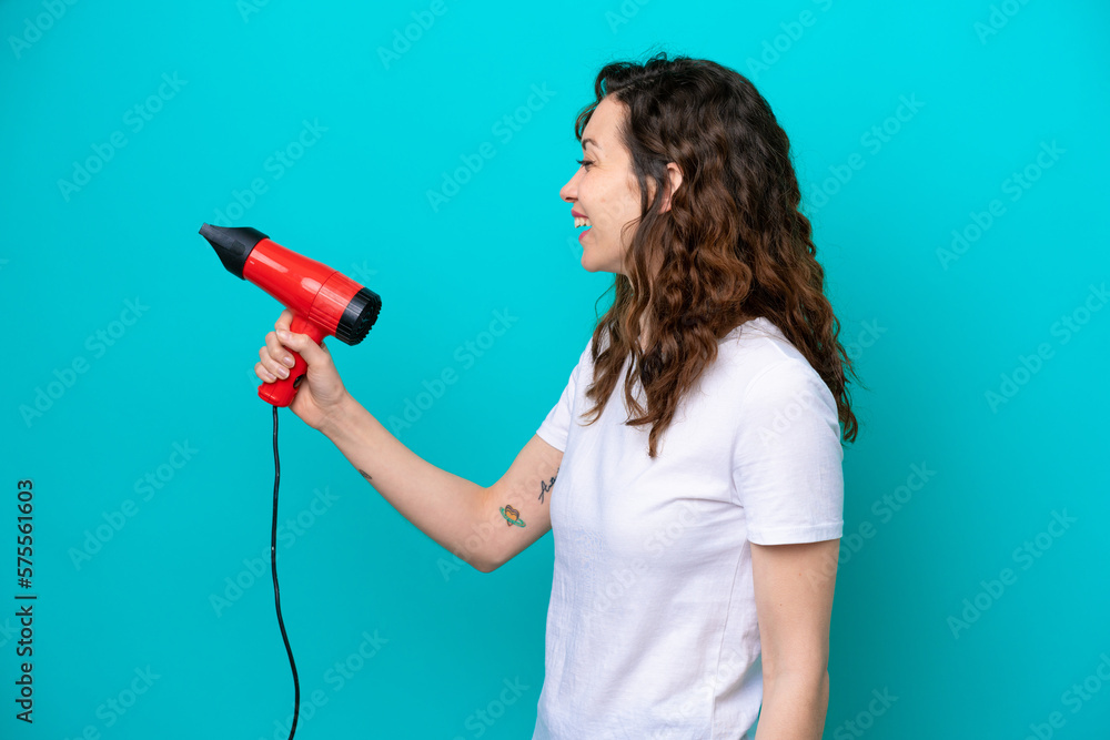 Young caucasian woman holding a hairdryer isolated on blue background with happy expression Young caucasian woman holding a hairdryer isolated on blue background with happy expression