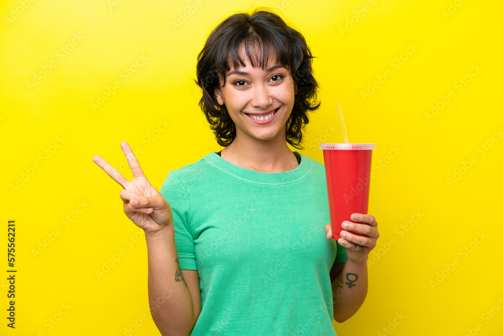 Young Argentinian woman holding a soda isolated on yellow background smiling and showing victory sign