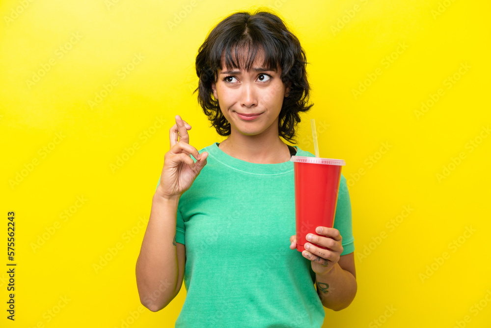 Young Argentinian woman holding a soda isolated on yellow background with fingers crossing and wishing the best Young Argentinian woman holding a soda isolated on yellow background with fingers crossing and wishing the best