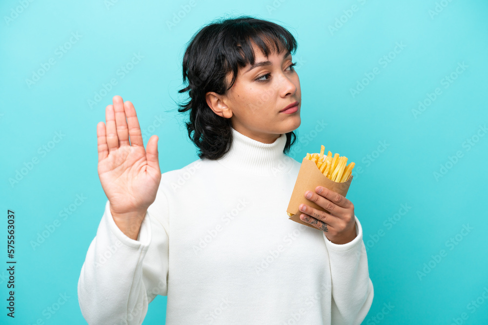 Young Argentinian woman holding fried chips isolated on blue background making stop gesture and disappointed Young Argentinian woman holding fried chips isolated on blue background making stop gesture and disappointed