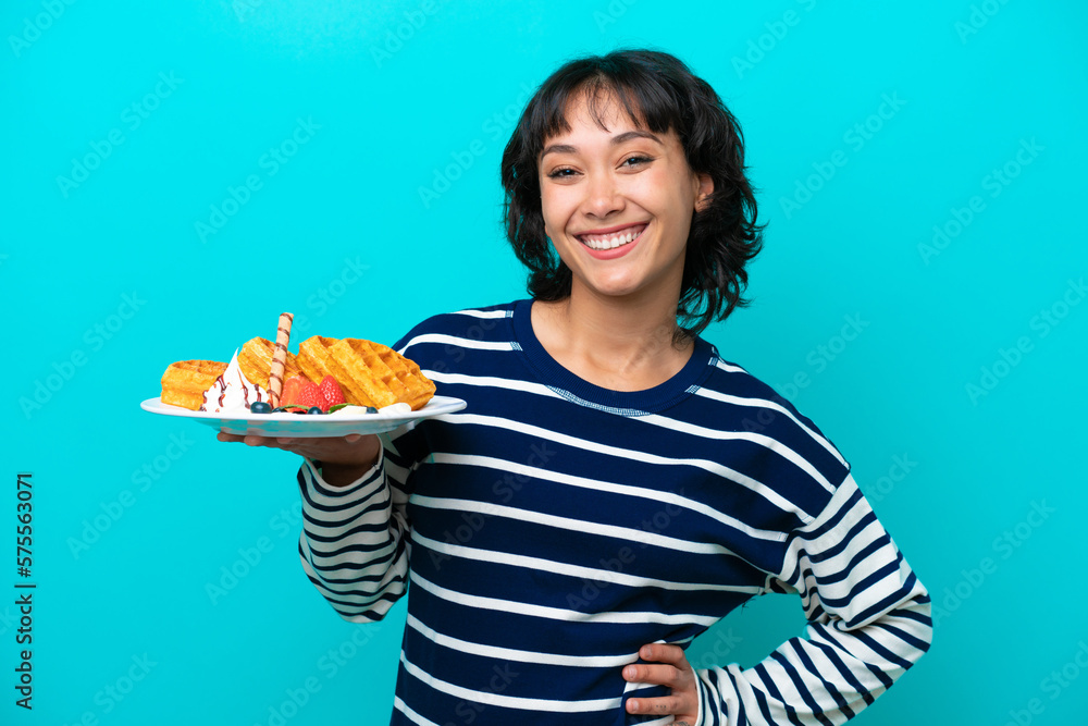 Young Argentinian woman holding waffles isolated on blue background posing with arms at hip and smiling