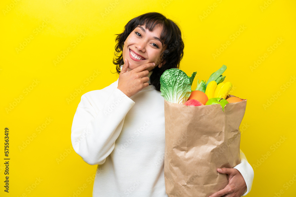 Young Argentinian woman holding a grocery shopping bag isolated on yellow background happy and smiling Young Argentinian woman holding a grocery shopping bag isolated on yellow background happy and smiling