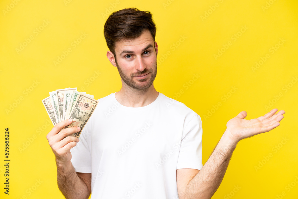 Young caucasian man taking a lot of money isolated on yellow background extending hands to the side for inviting to come Young caucasian man taking a lot of money isolated on yellow background extending hands to the side for inviting to come