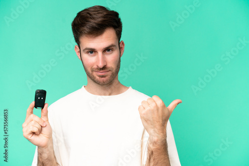 Young caucasian man holding car keys isolated on green background pointing to the side to present a product