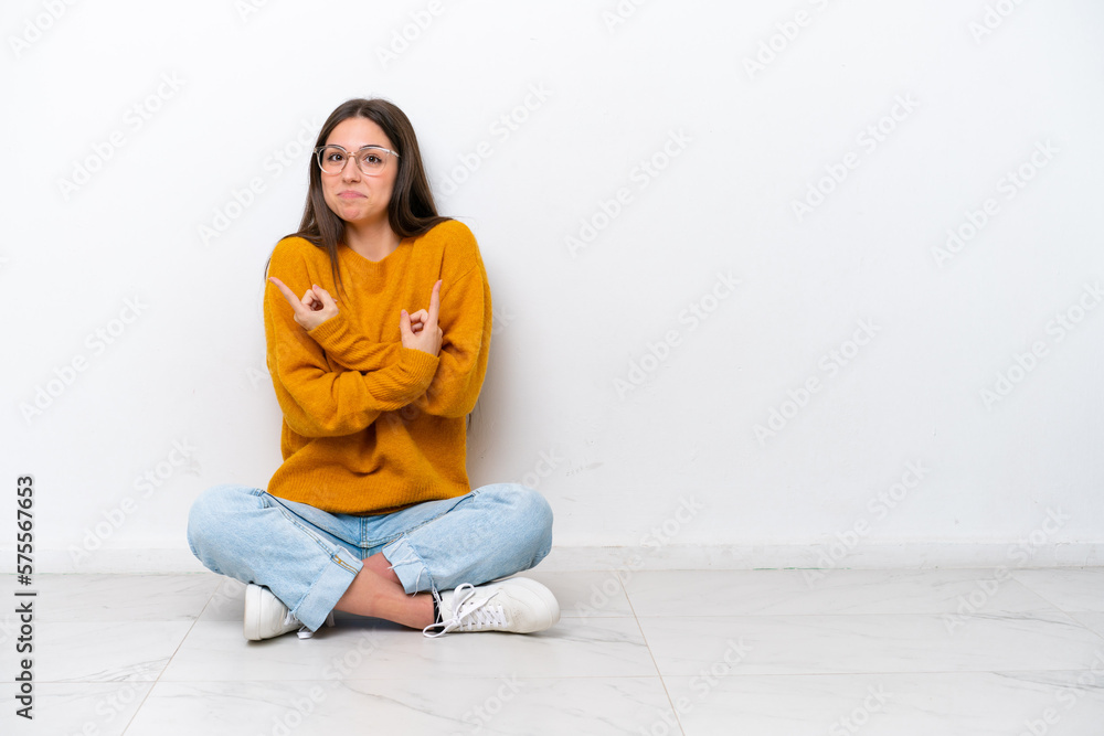 Young girl sitting on the floor isolated on white background pointing to the laterals having doubts Young girl sitting on the floor isolated on white background pointing to the laterals having doubts