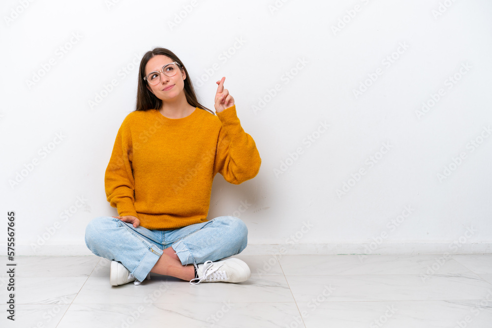 Young girl sitting on the floor isolated on white background with fingers crossing and wishing the best Young girl sitting on the floor isolated on white background with fingers crossing and wishing the best