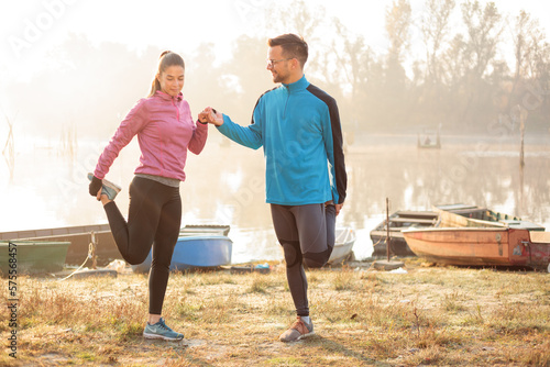 Beautiful young man and woman exercising, stretching in the early morning mist on the lake or river banks. Worming up for jogging. Healthy and active sport lifestyle