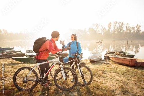 Smiling young man and woman riding mountain bikes by the lake or river, talking and resting. Sun rising through the mist above water in the background. Healthy and active lifestyle concept.
