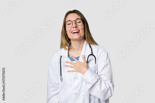 Compassionate female physician with a stethoscope around her neck  ready to diagnose and care for her patients in her signature white coat laughs out loudly keeping hand on chest.