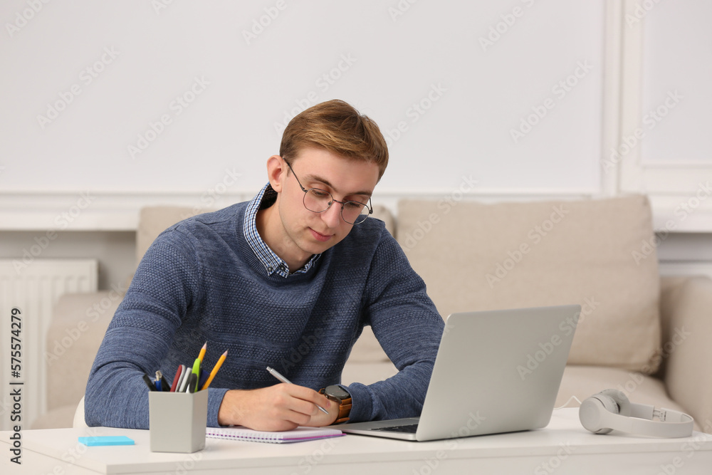 Young man with laptop writing in notebook at table indoors