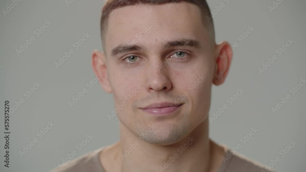 The young man looks up and starts smiling while looking straight into the camera. Portrait shot of a smiling man in front of a white wall. The young man looks up and starts smiling while looking straight into the camera. Portrait shot of a smiling man in front of a white wall.