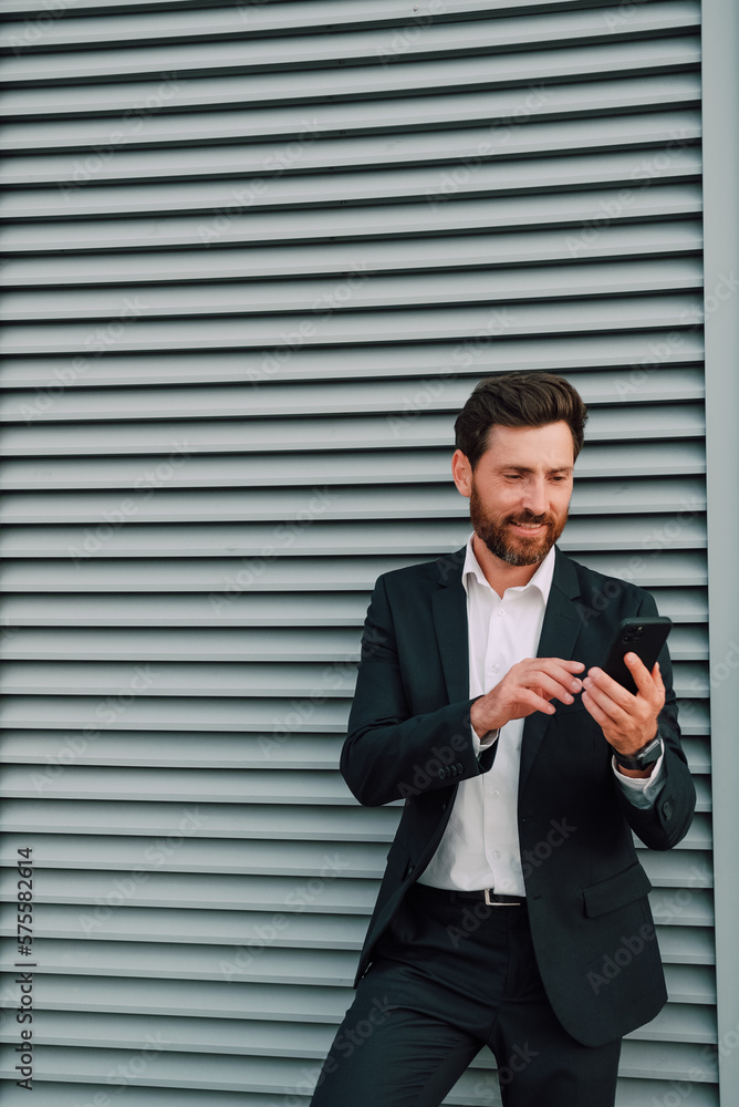 Businessman texting phone app in city street on brick wall background. Business man holding smartphone in suit. Urban young professional lifestyle. Businessman texting phone app in city street on brick wall background. Business man holding smartphone in suit. Urban young professional lifestyle.
