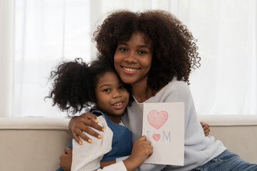 African American young mother hugging her chid daughter and holding mother day paper card at home