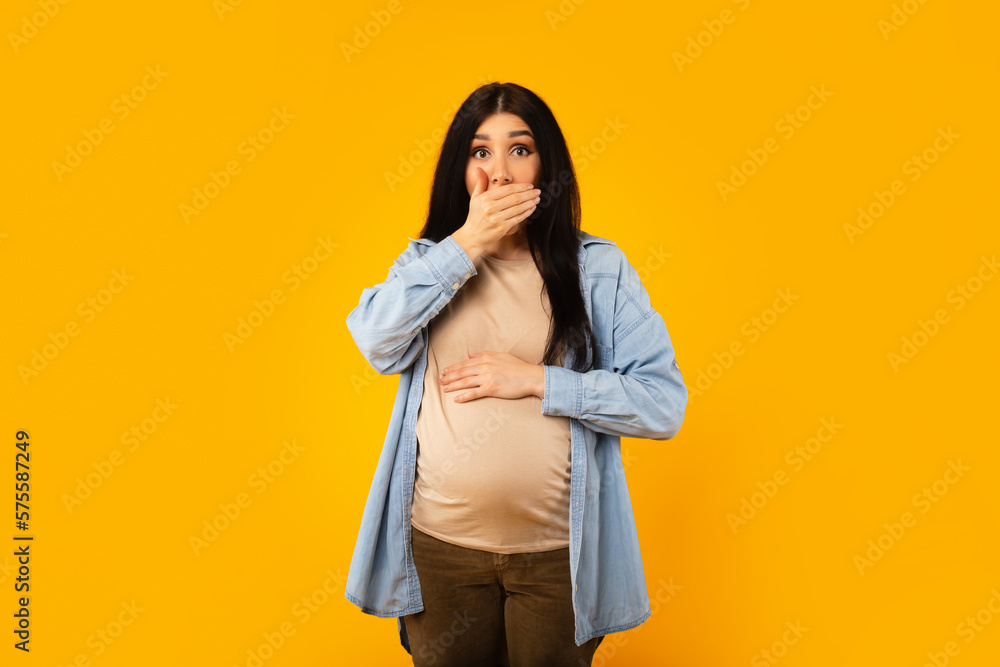 Portrait of young pregnant lady suffering nausea, covering mouth with hand, feeling unwell, yellow background Portrait of young pregnant lady suffering nausea, covering mouth with hand, feeling unwell, yellow background