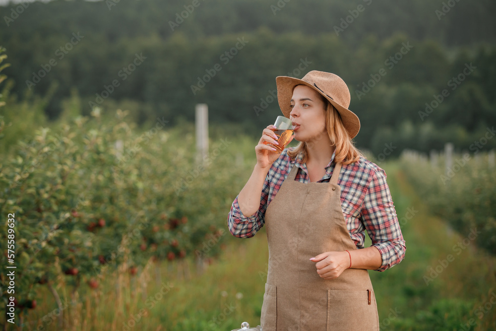 Happy smiling female farmer worker drinking tasty apple juice in glass standing in orchard garden during autumn harvest. Harvesting time