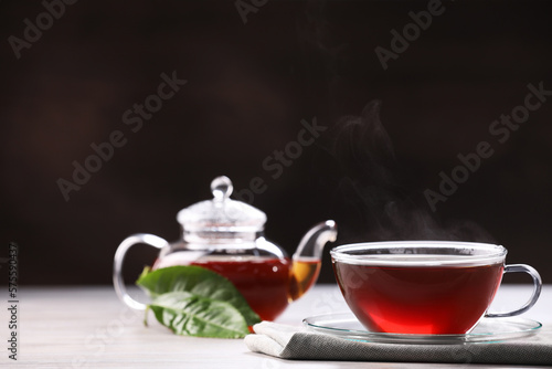 Aromatic hot tea in glass cup, teapot and leaves on white wooden table against black background. Space for text