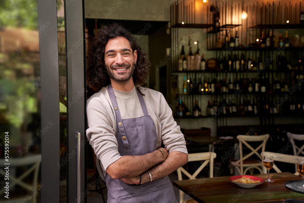 Portrait of happy young adult local small business owner looking at camera. Happy male bartender crossing his arms with positive attitude. Cheerful worker smiles while standing leaning on bar door. - obrazy, fototapety, plakaty 