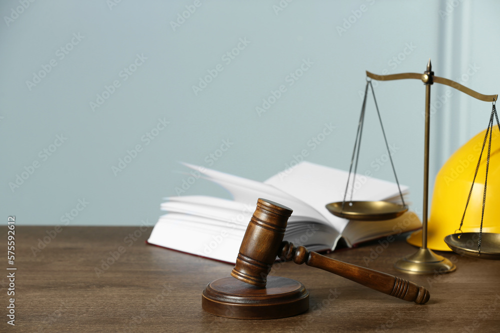 Labour, construction and land law concepts. Judge gavel, scales of justice, open book with protective helmet on wooden table, space for text Labour, construction and land law concepts. Judge gavel, scales of justice, open book with protective helmet on wooden table, space for text