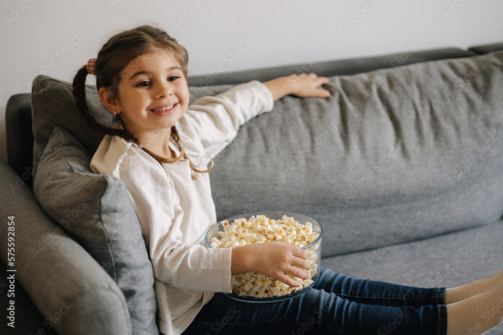 Adorable little girl watching TV at home and laughs. Cute girl eating popcorn. Holiday mood