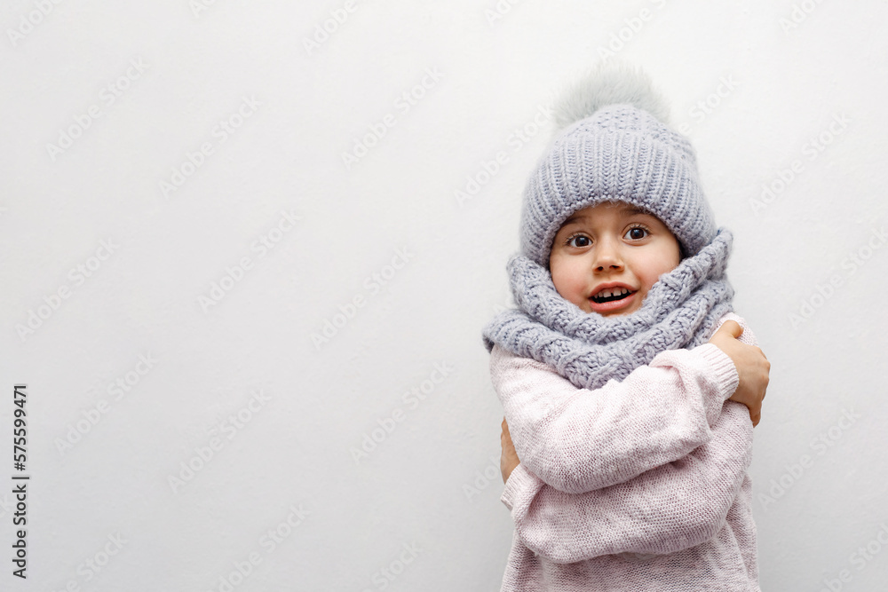 child girl in a gray hat and scarf hugs herself on a white background and smiles, grimace, childhood, winter season child girl in a gray hat and scarf hugs herself on a white background and smiles, grimace, childhood, winter season