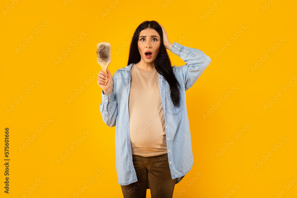 Lose hair during pregnancy. Shocked young pregnant lady holding comb in her hand and touching head, yellow background Lose hair during pregnancy. Shocked young pregnant lady holding comb in her hand and touching head, yellow background