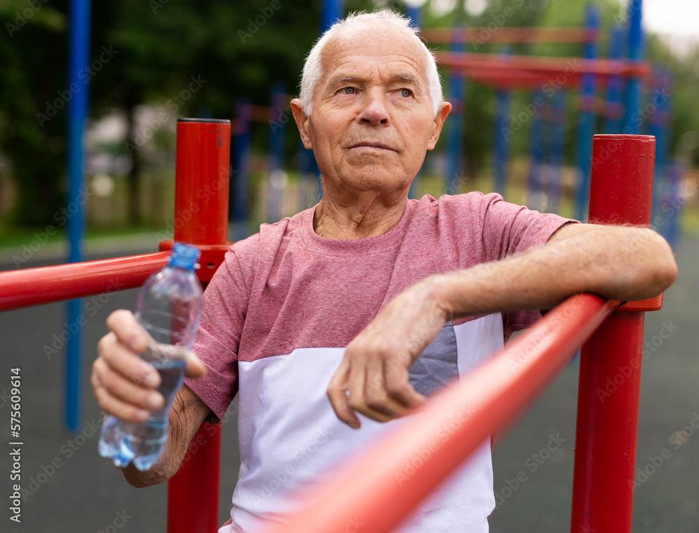 Obraz premium Senior man resting and drinking water from bottle after jogging in park