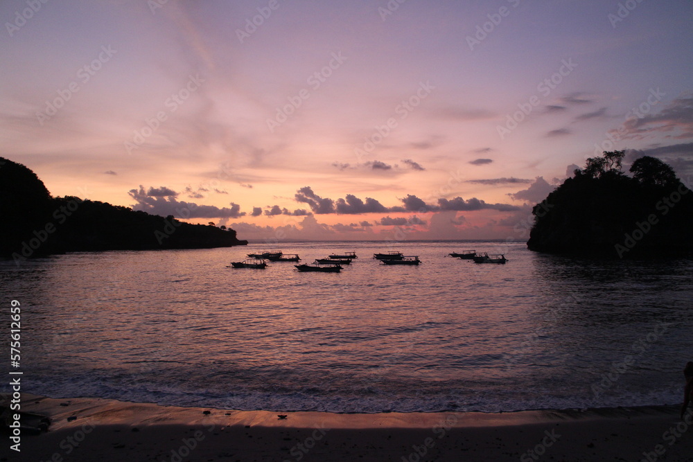 Colorful sunset in Nusa Penida, Bali. Purple, pink and orange colors. Water, boats and islands. Sunset reflexion in the ocean. Sun shine through the clouds. Island holidays, perfect evening. 