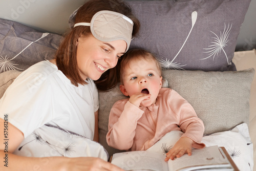 Horizontal shot of beautiful young adult young mother with her little daughter reading book in bed, expressing happiness, reads before go to sleep for charming infant kid.