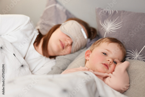 Portrait of dark haired female wearing blindfold lying with her baby in bed, daughter already wake up and looking around, mommy sleeping. Happy motherhood and childhood.