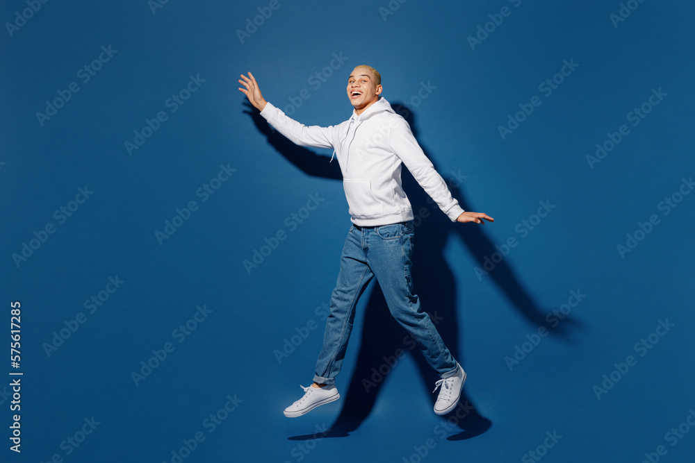 Full body young fun dyed blond man of African American ethnicity wear white hoody jump high pov flying in air with outstretched hands isolated on plain dark royal navy blue background studio portrait.