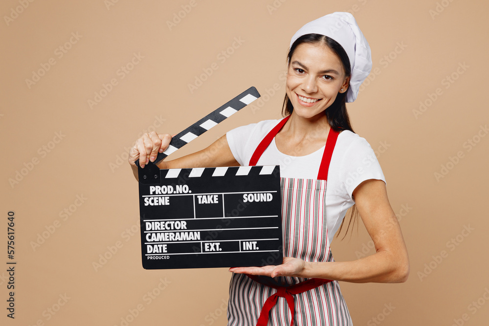 Young housewife housekeeper chef baker latin woman wear striped apron toque hat hold in hand classic black film making clapperboard isolated on plain pastel light beige background. Cook food concept.