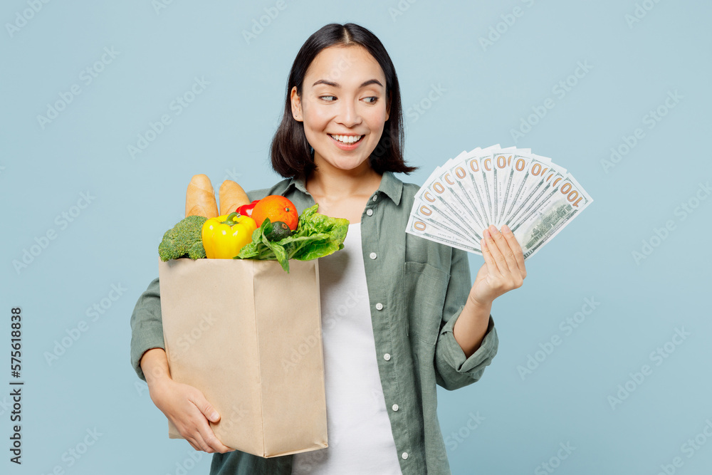 Young woman in casual clothes hold brown paper bag with food product fan of cash money in dollar banknotes isolated on plain blue background studio portrait. Delivery service from shop or restaurant. Young woman in casual clothes hold brown paper bag with food product fan of cash money in dollar banknotes isolated on plain blue background studio portrait. Delivery service from shop or restaurant.