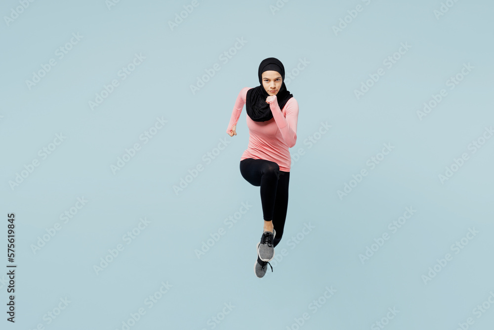 Full body young asian muslim fitness trainer sporty woman wear pink abaya hijab spend time in home gym running fast jump high isolated on plain blue background studio. Workout sport fit abs concept.