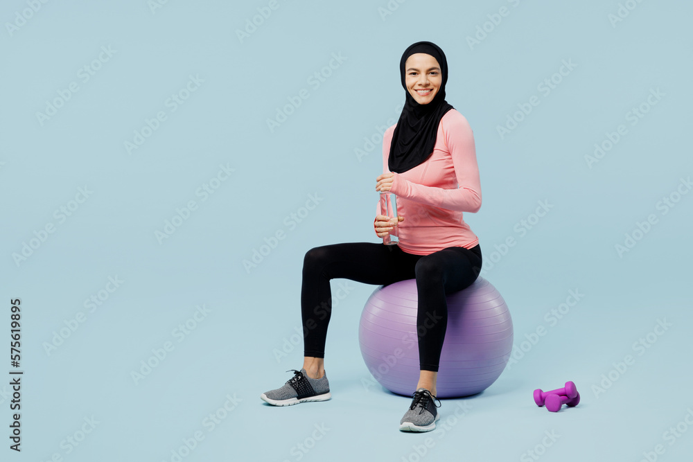 Full body young asian muslim fitness trainer sporty woman wear pink abaya hijab spend time in home gym sit fitball drink water isolated on plain blue background studio Workout sport fit abs concept Full body young asian muslim fitness trainer sporty woman wear pink abaya hijab spend time in home gym sit fitball drink water isolated on plain blue background studio Workout sport fit abs concept