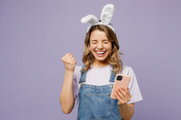 Young fun woman wearing casual clothes bunny rabbit ears hold in hand use mobile cell phone do winner gesture isolated on plain pastel light purple background studio. Lifestyle Happy Easter concept.