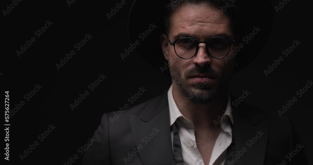 cool mysterious man in elegant tuxedo with open collar shirt and hat looking to side, adjusting suit and posing with flash lights on cool mysterious man in elegant tuxedo with open collar shirt and hat looking to side, adjusting suit and posing with flash lights on