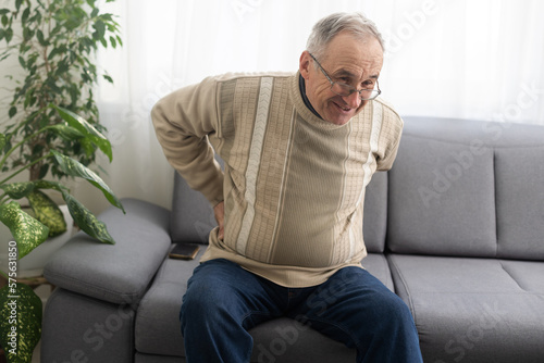 Caucasian old senior elderly unhealthy sickness grey male grandpa near sofa at home alone holding hands on back having emergency painful muscle and backache injury problem.