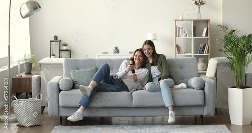 Cheerful mature mom with teen daughter jumped on couch, turn on TV, choose program with remote control, watch movies or telecast through online digital streaming services. Happy family leisure at home