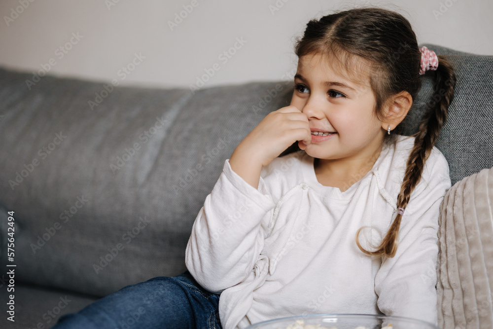 Adorable little girl watching TV at home and laughs. Cute girl eating popcorn. Holiday mood Adorable little girl watching TV at home and laughs. Cute girl eating popcorn. Holiday mood
